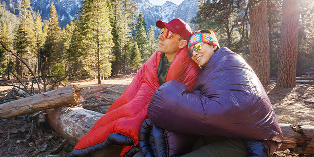 a photo of a man and a women wrapped up in re and purple sleeping quilts outside in nature